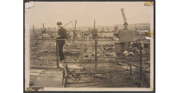 A black-and-white photograph of a White man standing among the ruins of a village.