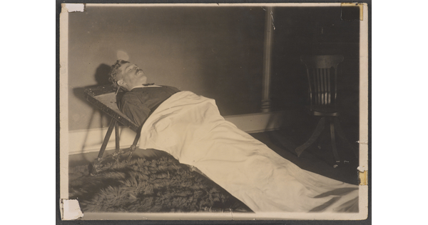 A black-and-white photograph of a White man in a formal suit laying on a stretcher and partially covered by a white sheet.