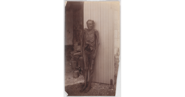 A black-and-white photograph of a mummified person standing upright and leaning against a wall.