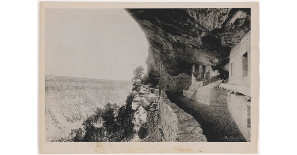 A black-and-white photograph of human-made structures built into a steep cliff face overlooking a deep valley.