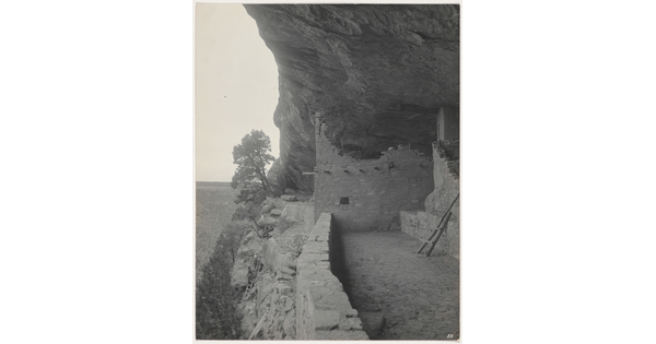 A black-and-white photograph of a dirt path next to human-made structures built under a cliff overhang.