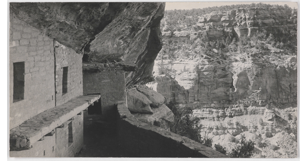 A black-and-white photograph of human-made stone structures in a cliff face overlooking a cliff wall across a valley.