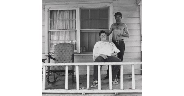 A black-and-white photograph of two White men on the porch of a house: one seated with a white apron around his neck and shoulders; the other standing, shirtless, holding clippers in one hand, the other on the seated man's head.
