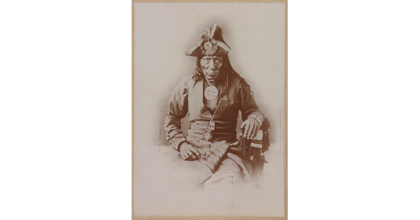 A black-and-white portrait photograph of a seated Indigenous man seated wearing a colonial-style jacket and a tricorn hat.