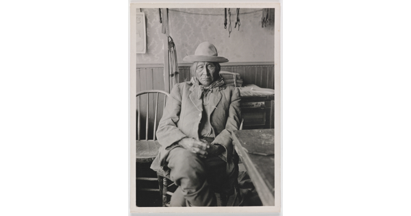 A black-and-white photograph of an Indigenous man in a rumpled suit seated next to a table with his hands in his lap.