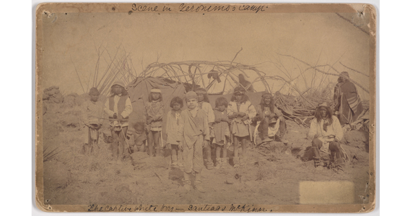 A slightly faded sepia-toned photograph of a group of Indigenous adults and children and one White child standing in front of a camp.