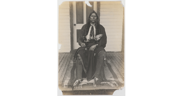 A black-and-white photograph of an Indigenous man with long braids seated on a wooden porch.