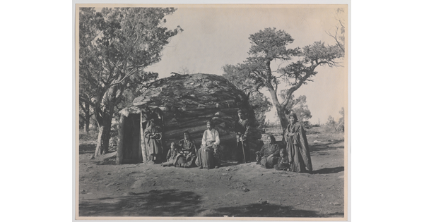 A black-and-white photograph of a group of Indigenous adults and children in front of a round, wood dwelling.