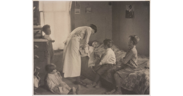 A black-and-white photograph of a nurse bending over a person in a bed, two children sitting on the bed, and two children behind the nurse.
