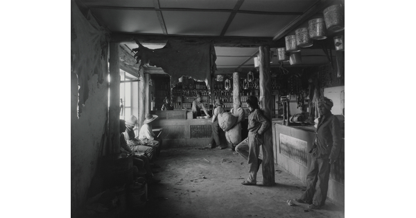 A black-and-white photograph of Indigenous people in a shop with a variety of products such as animal hides, tin pails, and lanterns hanging from the ceiling.