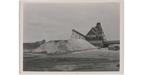 A black-and-white photograph of large pile of salt in front of a multi-story industrial building with a sign that reads "Crystal Salt Co."