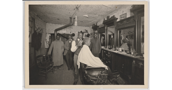 A black-and-white snapshot of the inside of a barber shop with a Black man in a chair getting his hair cut.
