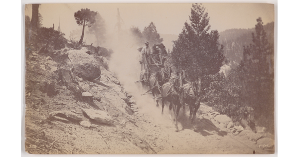 A black-and-white photograph of a stagecoach being pulled by six horses down a dusty dirt road on a hillside.