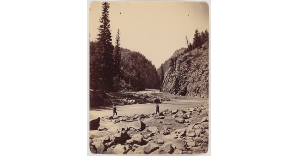 A black-and-white photograph of two men fishing in a river surrounded by a rocky shore and cliffs.