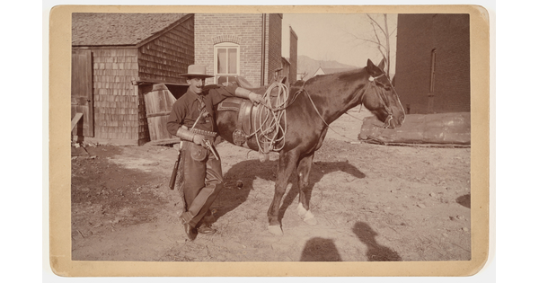 A black-and-white photograph of a light-skinned man with a large mustache, wearing a cowboy hat, holding a gun and leaning against a horse.