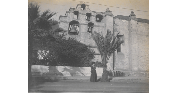 A black-and-white photograph of a woman standing next to a building with several large bells hanging near the top.