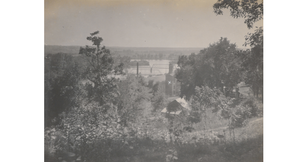 A black-and-white photograph taken from a higher elevation of a bridge spanning a river and forested area that includes some buildings.