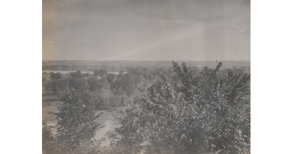 A black-and-white photograph taken from a higher elevation of a forested area and a river in the distance.