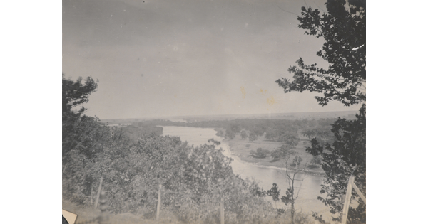 A black-and-white photograph taken from a higher elevation of a river and surrounding fields and trees.