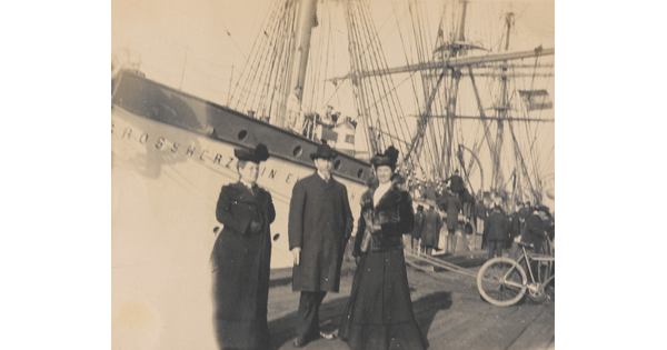 A black-and-white photograph of three nicely dressed adults standing on a dock in front of a massive ship.