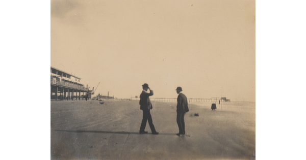 A black-and-white photograph of two men in bowler hats and suits standing on a sandy beach.