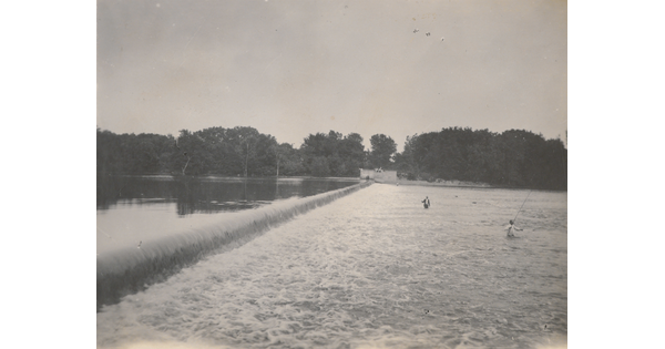 A black-and-white photograph of people fishing in a rushing river near a dam.
