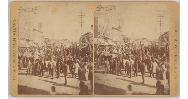 A sepia-toned stereograph of a large gathering of people in a street lined with buildings.