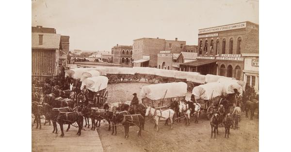 A sepia-toned photograph of covered wagons arranged in a large circle on a dirt street in a town.