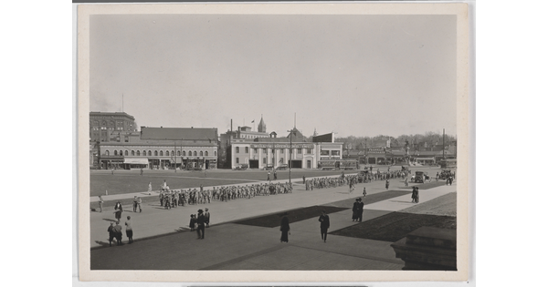 A black-and-white photograph of a parade coming through a wide street as scattered spectators watch.