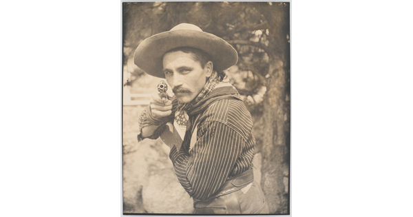 A sepia-toned photograph of a White man in a cowboy hat and bandana pointing a revolver at the camera.