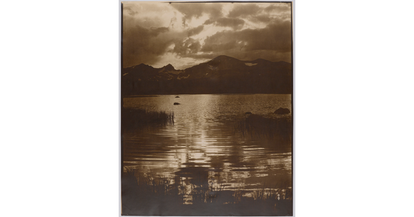 A sepia-toned photograph of light filtered through clouds reflecting on a lake surrounded by mountains.