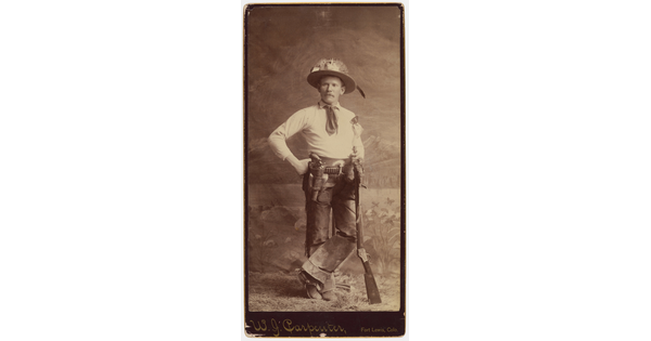 A sepia-toned studio portrait photograph of a cowboy wearing a hat decorated with feathers, a belt holding a revolver, and leaning on the muzzle of a rifle.
