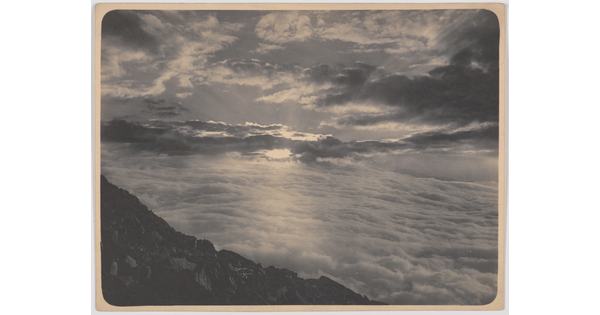 A black-and-white photograph of a dramatic cloudscape with the sun and rays peeking through below a rocky landscape in the foreground.