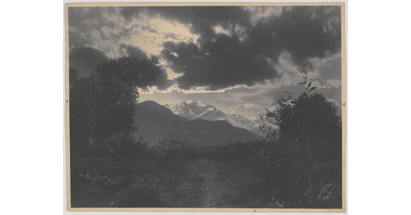 A black-and-white photograph of snow-capped mountains under dark and sun-lit clouds framed by trees.