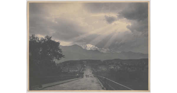 A black-and-white photograph of two people walking down a road toward a town; snow-capped mountain and sun rays in the distance.