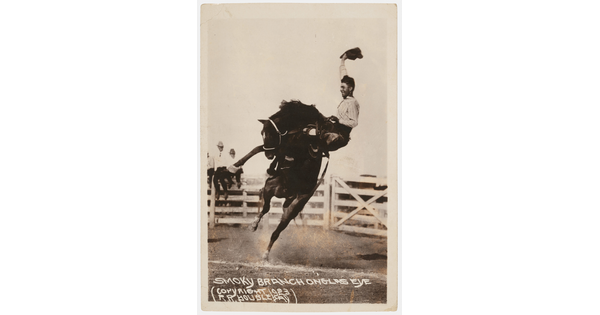 A black-and-white photograph of a cowboy waving his hat as he holds on to a rearing horse at an outdoor rodeo.