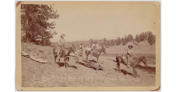 A sepia-toned photograph of several cowboys on horseback hauling large pieces of wood with ropes.
