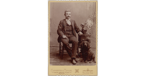 A black-and-white portrait photograph of a seated White man with a mustache wearing a three-piece suit and a dog sitting at his feet.