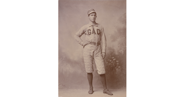 A sepia-toned portrait photograph of a White man, one hand on hip, wearing a baseball hat and uniform with "GAC" across the chest.