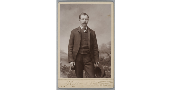 A black-and-white studio photograph of a young White man with a mustache wearing a three piece suit, holding a hat in one hand.