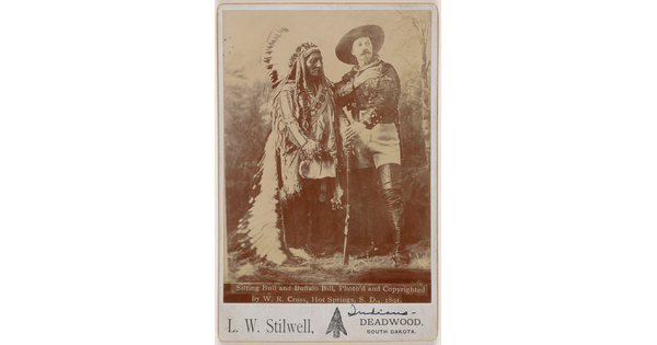A sepia-toned portrait photograph of an Indigenous man wearing a feathered headdress and traditional clothing next to a White man in a large hat and tall leather boots holding a rifle in front of them.