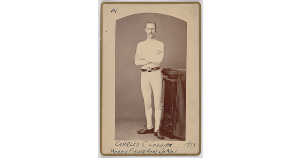 A black-and-white portrait photograph of a young White man with a mustache wearing all white standing with arms crossed next to a decorative column.