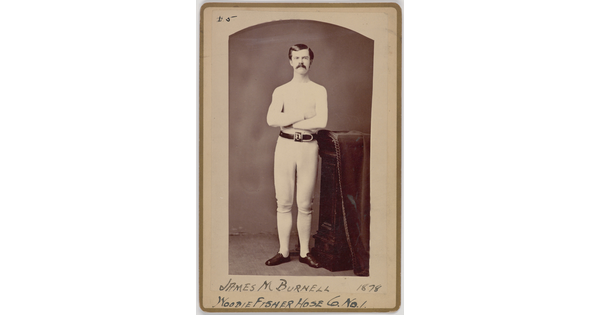 A black-and-white portrait photograph of a young White man with a mustache wearing all white standing with arms crossed next to a decorative column.
