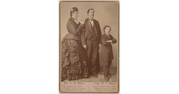 A sepia-toned portrait photography of a very tall, White woman in an elaborately draped dress next to a very tall, White man, and a White man of average height, both in suits.