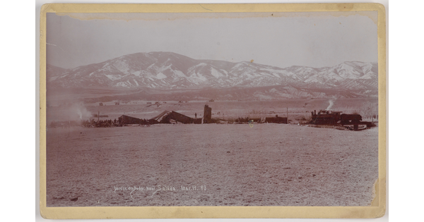 A black-and-white photograph of mangled train cars and an engine on a plain with snow-covered mountains in the distance.