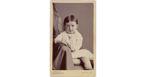 A black-and-white portrait photograph of a formally-dressed White child posed on a chair draped in fabric.