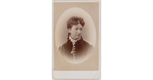 A black-and-white oval-shaped portrait photograph of a White woman, hair pulled up with ringlets that lay on her shoulders, wearing a dress with a high ruffled neck.