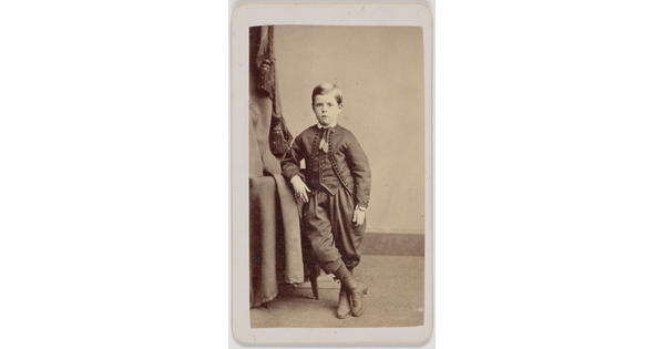 A black-and-white portrait photograph of a White boy wearing a formal jacket and vest over knickerbocker pants, leaning on a table, one leg crossed over the other.