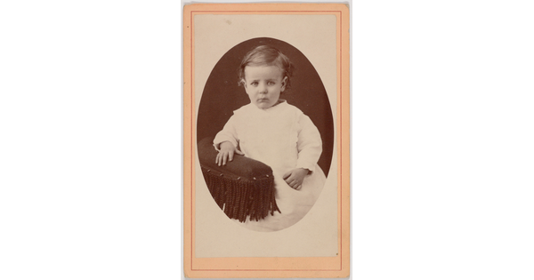 A black-and-white oval-shaped portrait photograph of a White toddler in a white dress resting one hand on the fringed arm of a chair.