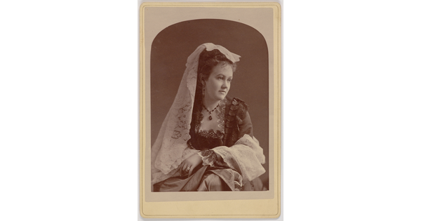 A black-and-white portrait photograph of a young White woman with a white lace veil on her head, wearing a dress with a square neckline trimmed in lace, her arms crossed on her lap.